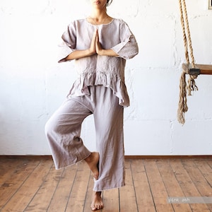 May include: A two-piece set in a muted pink ash colour, consisting of a loose-fitting top with elbow-length sleeves and a ruffled hem, paired with matching wide-leg trousers. The garment features a raw-edge finish at the neckline and cuffs.