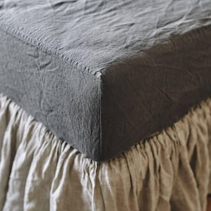 May include: A charcoal gray fitted sheet with a textured, wrinkled appearance, featuring a structured corner seam. The sheet is paired with a light beige bed skirt that has a gathered, ruffled design along the bottom edge of the mattress.