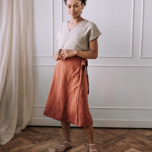May include: A woman wearing a light beige linen top and a rust-colored linen wrap skirt. She is standing on a wood floor with a white wall and a white curtain in the background.