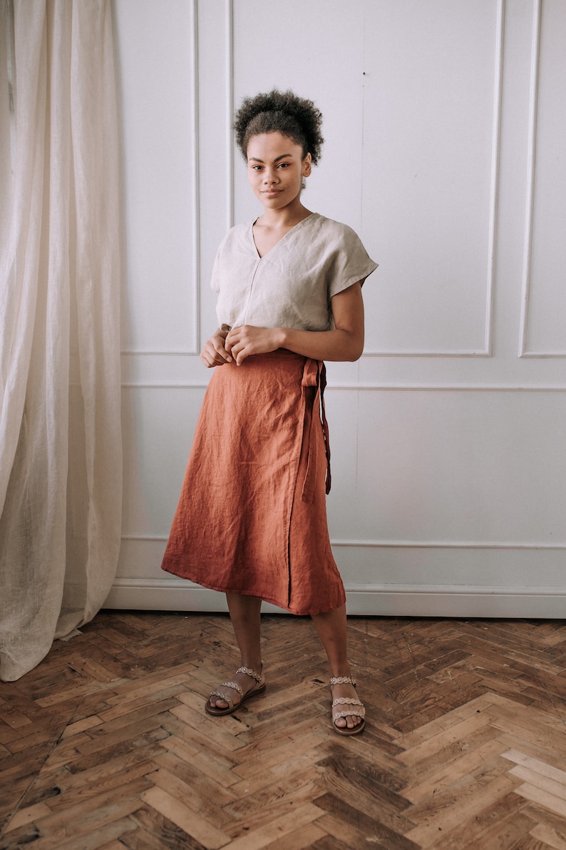May include: A woman wearing a light beige linen top and a rust-colored linen wrap skirt. She is standing on a wood floor with a white wall and a white curtain in the background.
