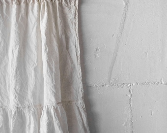 May include: An antique white curtain featuring a gathered top header and a horizontal ruffled hem detail. The fabric has a visible texture and hangs in vertical folds, displaying a soft, neutral tone throughout the entire piece.