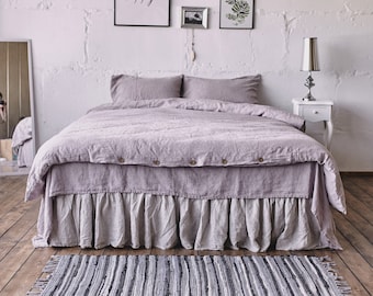 May include: A light purple linen duvet cover with a ruffled bed skirt and a matching set of pillowcases. The bed is made with a gray and white striped rug on a wooden floor. The wall behind the bed is white brick with a framed print that reads "Think happy. Be happy."