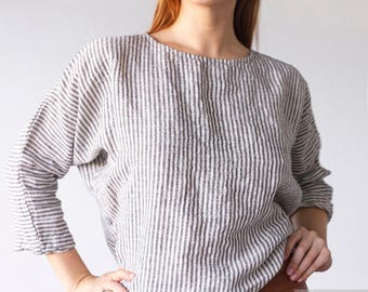 May include: A long-sleeved top featuring a vertical striped pattern in charcoal gray and off-white. The garment has a rounded neckline, a relaxed fit, and three-quarter length sleeves. The fabric appears to have a textured, woven finish.