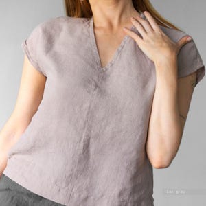 May include: A flax gray short-sleeved top featuring a V-neckline and a visible woven texture. The garment has a relaxed fit with cuffed sleeves and a straight hemline. The fabric appears to be a natural fiber with a subtle, earthy tone.