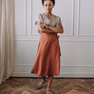 May include: A woman wearing a light brown linen top and a rust-colored linen wrap skirt. She is standing in front of a white wall with a white trim. The floor is a herringbone pattern of wood.