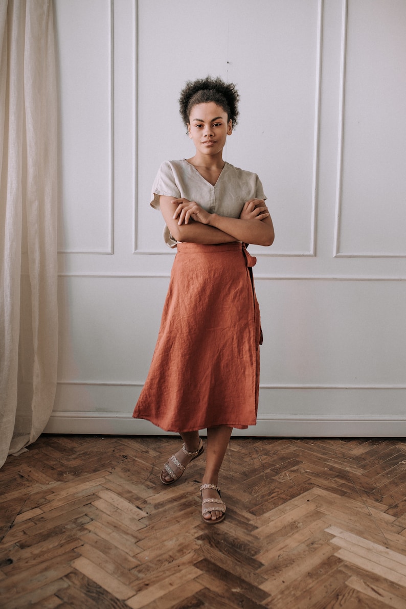 May include: A woman wearing a light brown linen top and a rust-colored linen wrap skirt. She is standing in front of a white wall with a white trim. The floor is a herringbone pattern of wood.