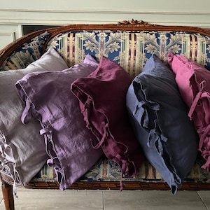 Può includere: Un set di cinque cuscini quadrati in lino in varie tonalità di grigio chiaro, lavanda, prugna scuro, blu ardesia e rosa antico. Ogni cuscino presenta un bordo decorativo con laccetti in tessuto agli angoli e una trama visibile.