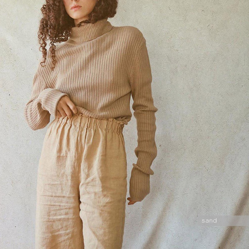 May include: A beige ribbed knit turtleneck sweater and matching linen pants. The long-sleeved sweater has a high neck and fitted cuffs. The pants have a gathered waistband. The word "sand" is in the bottom right corner.