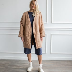May include: A beige quilted open-front coat with long sleeves and a relaxed fit, worn over a dark blue top and dark blue cropped trousers, paired with white lace-up boots. The coat features a subtle diamond-stitched pattern throughout the fabric.