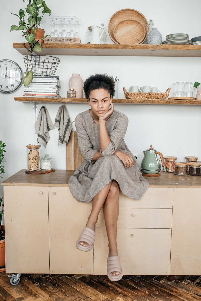 May include: A woman wearing a gray linen dress sits on a kitchen counter with her chin resting on her hand. She is wearing light pink slide sandals. The counter is made of wood and has two drawers. There are various kitchen items on the counter, including a green electric kettle, jars of spices, and a jar of cookies.