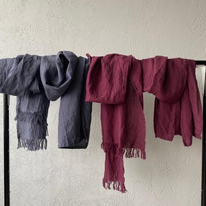 May include: Two long rectangular scarves draped over a black metal rack. One scarf is a muted slate gray color, and the other is a deep burgundy red. Both scarves feature a textured fabric appearance and have frayed, fringed edges at the ends.