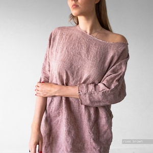 May include: A rose brown long-sleeved top with a wide, asymmetrical neckline that exposes one shoulder. The garment features a textured, woven fabric with raw, frayed edges along the neckline and sleeve cuffs, creating a loose, relaxed fit.