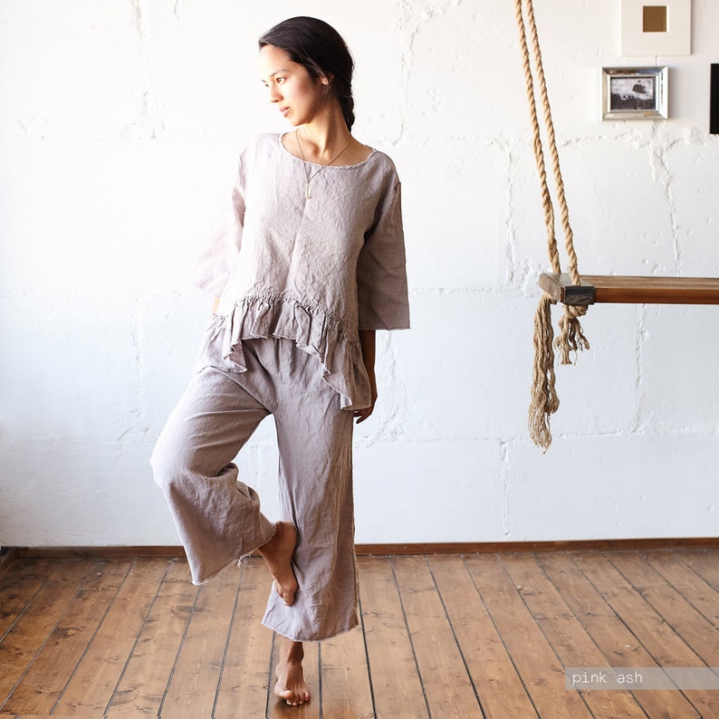 May include: A two-piece linen outfit in pink ash. The top has a round neckline and ruffled detail at the waist. The matching pants are wide-legged. The outfit is shown in a room with a wooden floor and a swing.