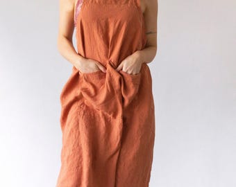 May include: A rust-colored apron featuring a square neckline, thick shoulder straps, and two large front pockets. The apron has a textured fabric appearance and falls to a mid-calf length, providing full coverage over the torso and legs.