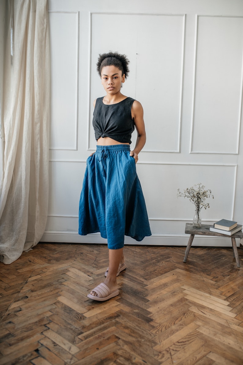 Pu&ograve; includere: Una persona indossa un top nero senza maniche con un nodo sul davanti e una gonna blu con tasche. La gonna &egrave; lunga fino al ginocchio. L'outfit &egrave; abbinato a sandali rosa. Lo sfondo comprende un muro bianco, un pavimento in legno e una tenda.