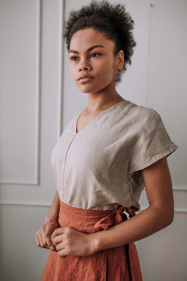 May include: A woman wearing a light brown linen top with a V-neck and short sleeves, paired with a rust-colored linen wrap skirt.