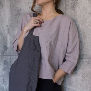 May include: A light purple linen top with a single pocket and three-quarter sleeves. A dark gray tote bag is slung over the shoulder. The person is wearing black pants.