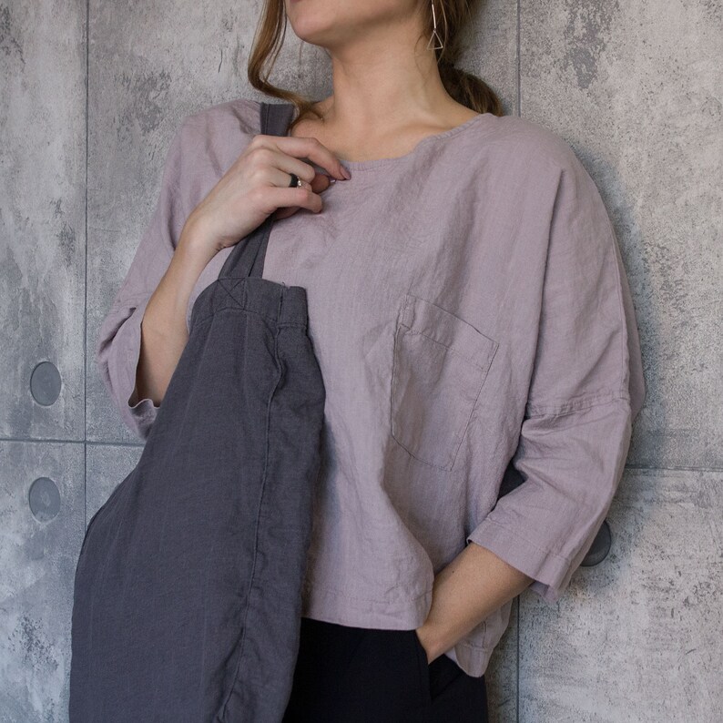 May include: A light purple linen top with a single pocket and three-quarter sleeves. A dark gray tote bag is slung over the shoulder. The person is wearing black pants.