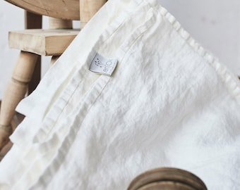 May include: An antique white textile with a visible woven texture and a stitched hem. A small white fabric label is attached to the corner, featuring a graphic of a plant and the text "lenok" in black lettering. The fabric is draped and folded.
