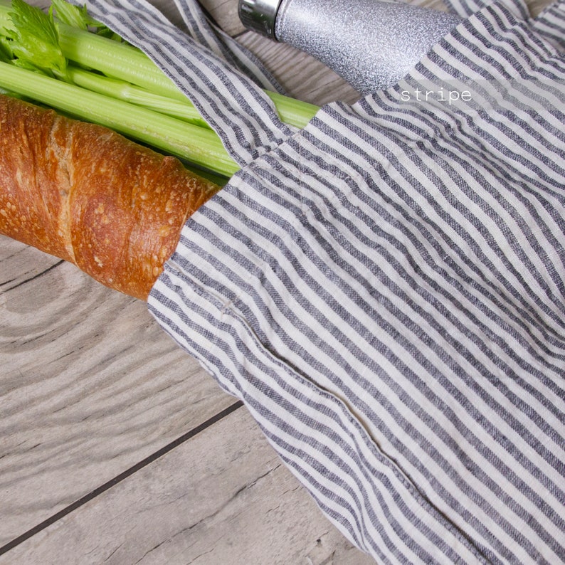 May include: A striped tote bag with a baguette, celery, and a silver water bottle. The bag is white with gray stripes and has long straps. The baguette is golden brown, and the celery is green.