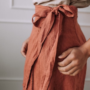 May include: A rust-colored linen wrap skirt with a self-tie sash. The skirt is tied at the waist and the sash is tied in a bow at the front.
