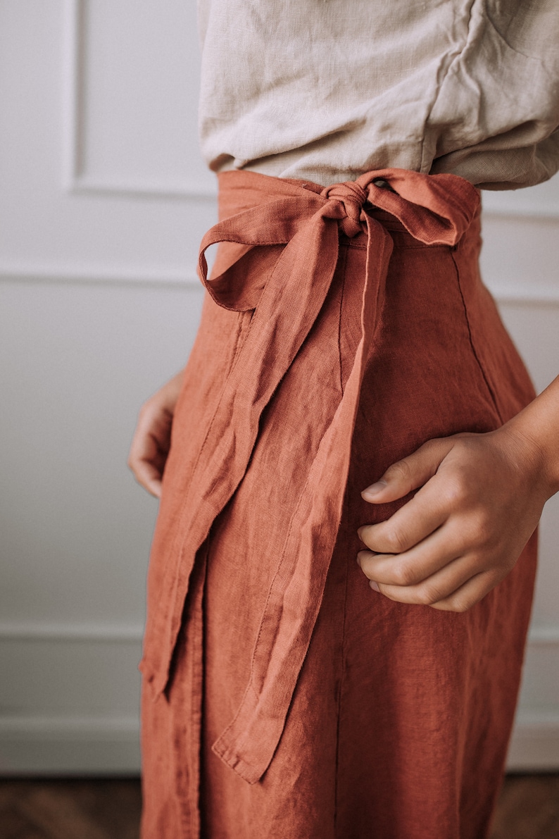 May include: A rust-colored linen wrap skirt with a self-tie sash. The skirt is tied at the waist and the sash is tied in a bow at the front.