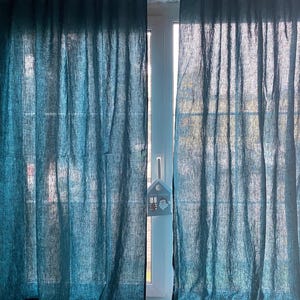 May include: Two teal blue curtains with a visible woven texture hanging from a rod with tab tops. The curtains are semi-sheer, allowing light to pass through, and feature a small house-shaped ornament hanging from the center window handle.