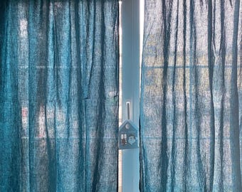 May include: Two teal blue curtains with a visible woven texture hanging from a rod with tab tops. The curtains are semi-sheer, allowing light to pass through, and feature a small house-shaped ornament hanging from the center window handle.