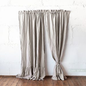 May include: A pair of flax gray curtains featuring a gathered ruffle at the top and a wide ruffled hem at the bottom. The fabric has a visible textured weave and a soft, draped appearance. One curtain panel is tied back with a matching fabric tie.