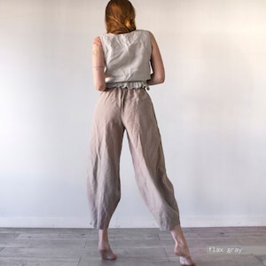 Pode incluir: Uma pessoa vestindo uma cal&ccedil;a de linho larga de cor cinza claro. A cal&ccedil;a tem um caimento solto e fluido e &eacute; cortada no tornozelo. A pessoa est&aacute; em p&eacute; em um piso de madeira.