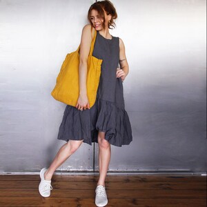 May include: A woman wearing a sleeveless gray dress with a ruffled hem and white sneakers. She is carrying a large, mustard-yellow tote bag. The background is a metallic silver wall and a wooden floor.