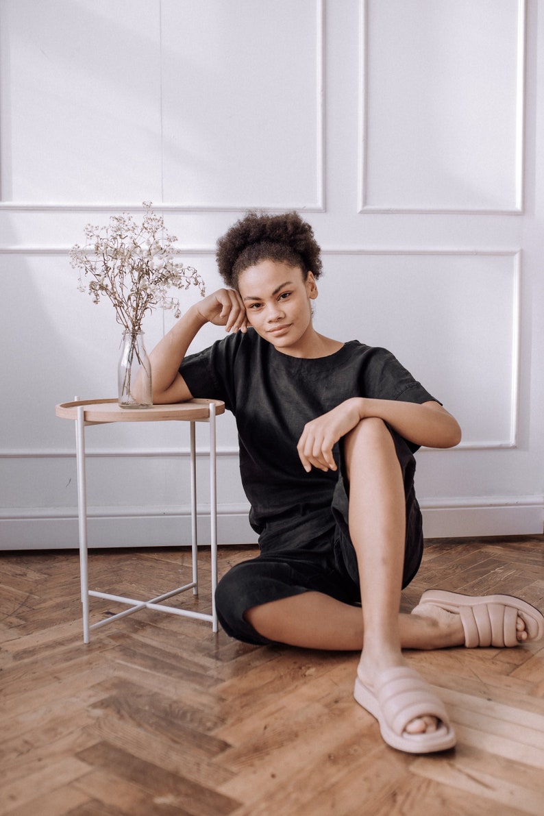Peut inclure: Une jeune femme aux cheveux boucl&eacute;s fonc&eacute;s est assise sur un sol en bois, v&ecirc;tue d'un t-shirt noir et d'un pantalon noir. Elle porte des tongs en fourrure rose clair. Une petite table blanche avec un vase de fleurs s&eacute;ch&eacute;es se trouve &agrave; c&ocirc;t&eacute; d'elle.