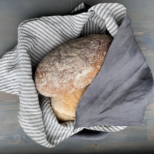 May include: Two loaves of bread wrapped in a reversible fabric cloth. One side of the cloth features a pattern of thin white and dark gray stripes, while the other side is a solid dark gray. The bread is partially covered by the folded fabric.