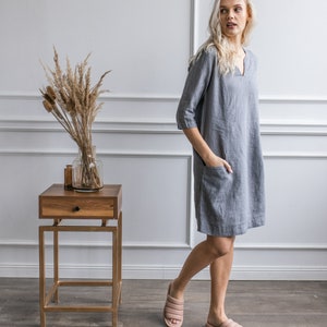 May include: A woman wearing a gray linen dress with a v-neck and pockets, standing in a room with white walls and a wooden side table. She is wearing light pink sandals.