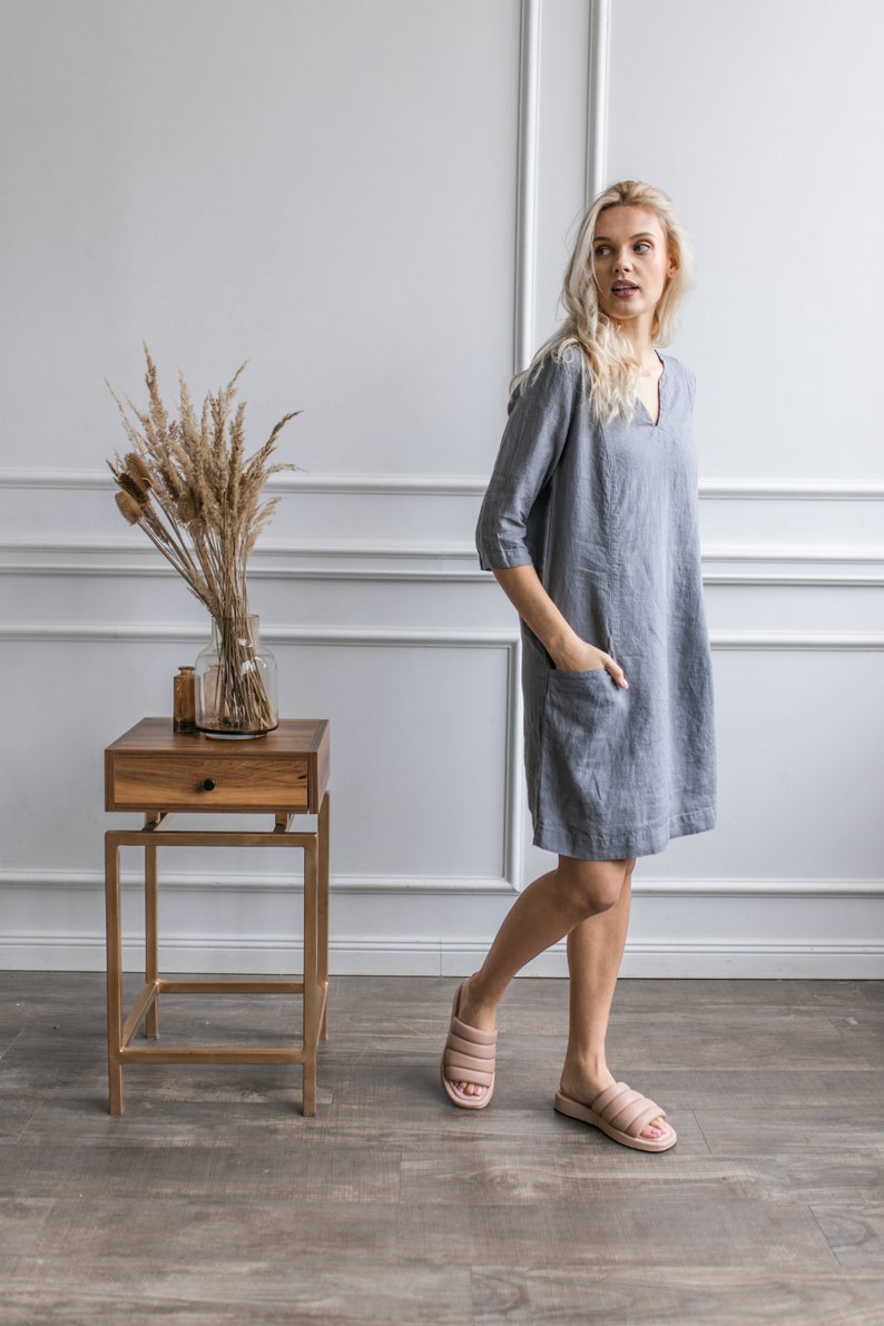 May include: A woman wearing a gray linen dress with a v-neck and pockets, standing in a room with white walls and a wooden side table. She is wearing light pink sandals.