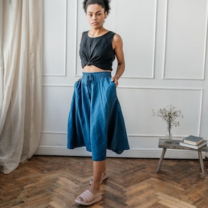 May include: A black sleeveless top with a knotted front hem paired with a calf-length blue skirt featuring an elastic waistband, a drawstring tie, and side pockets. The skirt has a loose, flowing silhouette and is made of a textured fabric.