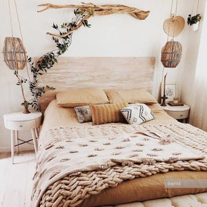 May include: A bed with a light wood headboard, tan bedding, and a chunky knit throw blanket with tassels. Two round white nightstands flank the bed, each with a hanging woven pendant light. A piece of driftwood with greenery is mounted above.