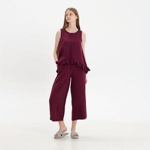 May include: A deep burgundy sleeveless top featuring a ruffled hemline paired with matching wide-leg cropped trousers. The set is made of a textured fabric and is worn with light gray cross-strap slide sandals on a plain white background.