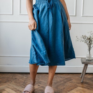 Pu&ograve; includere: Una persona indossa una gonna blu con coulisse in vita e un top nero. La gonna &egrave; lunga fino al ginocchio ed &egrave; realizzata in un tessuto fluido. Indossa anche sandali rosa. Lo sfondo &egrave; un muro bianco e un pavimento in legno.