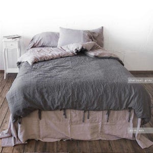 May include: A bed featuring a charcoal grey duvet cover and two matching pillows, layered over a pink ash bed skirt. The bedding has a textured, linen-like appearance. A white bedside table stands to the left of the bed on a wooden floor.