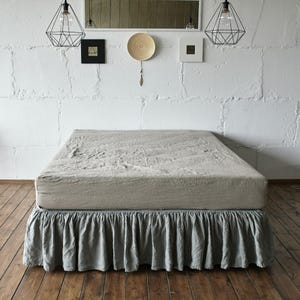 May include: A bed featuring a light beige, textured fabric bedspread and a matching light gray ruffled bed skirt. The bed is positioned against a white brick wall with two black geometric pendant lights hanging above the head of the bed.