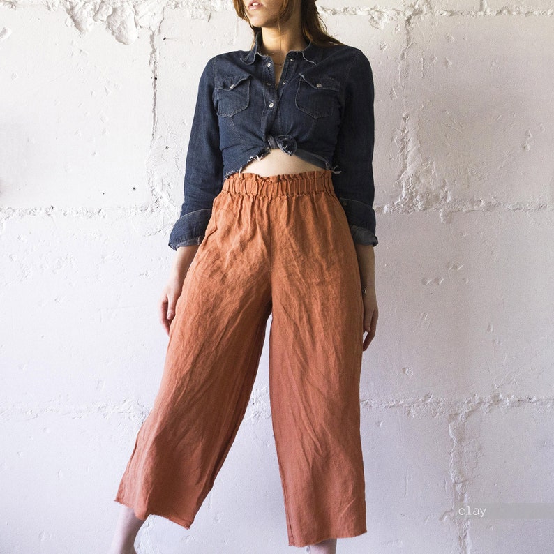 May include: Rust-colored linen culottes with an elastic waistband. The wide-leg pants are paired with a dark denim shirt tied at the waist. The outfit is set against a textured white wall, showcasing a casual, earthy style.