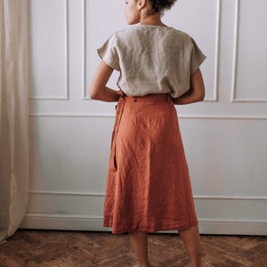 May include: A woman wearing a light brown linen top and a rust-colored linen wrap skirt. The skirt has a tie at the waist and falls to the mid-calf.