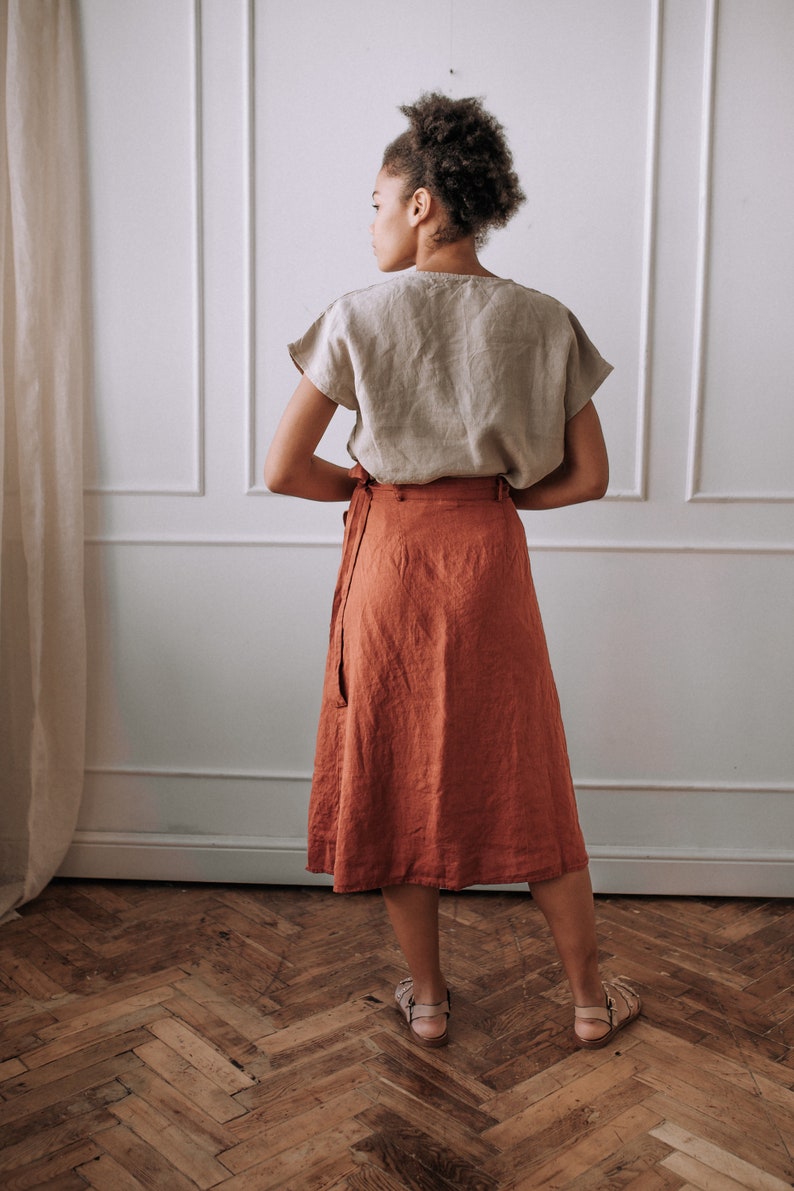 May include: A woman wearing a light brown linen top and a rust-colored linen wrap skirt. The skirt has a tie at the waist and falls to the mid-calf.