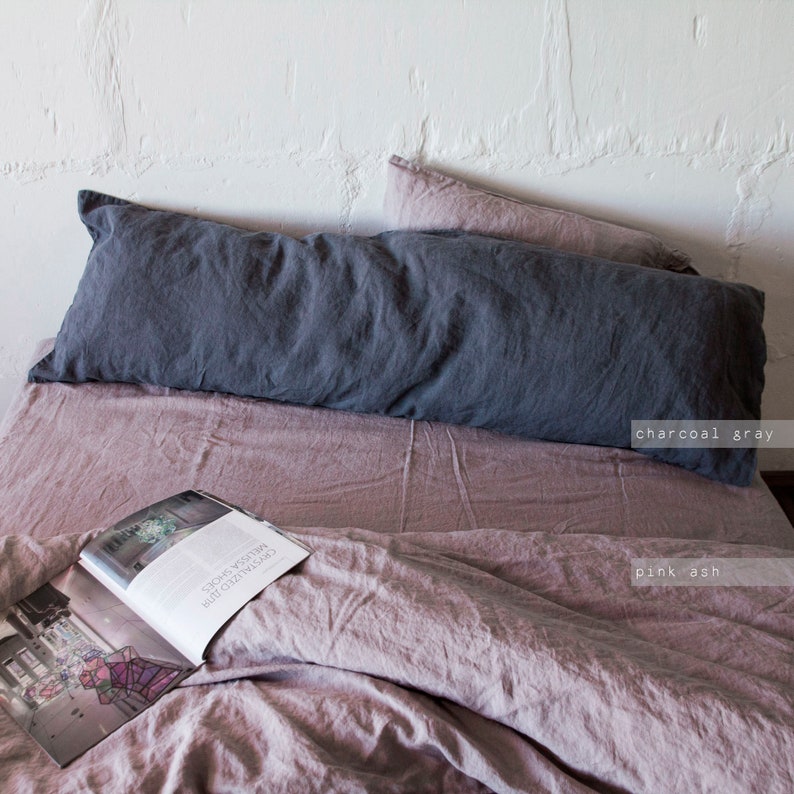 May include: A charcoal gray linen body pillow on a pink ash linen bed with a magazine on top of the bedding.