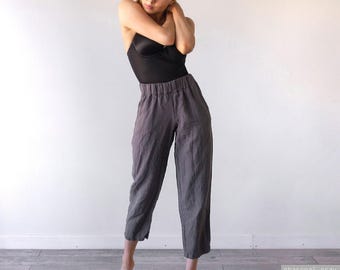 May include: Charcoal gray trousers featuring an elasticized waistband and a relaxed, cropped leg silhouette. The pants are crafted from a textured fabric and are paired with a black sleeveless bodysuit. The text charcoal gray is visible.