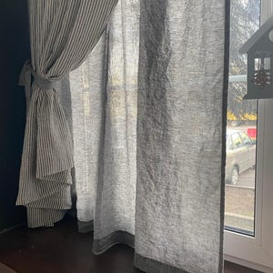 May include: A window curtain set featuring a grey and white striped panel tied back with a matching fabric tie, paired with a semi-sheer, textured grey panel. A small grey house-shaped ornament with the word Merry is hanging on the window frame.