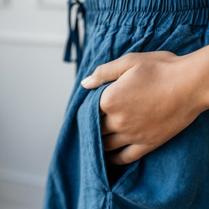 Pu&ograve; includere: Primo piano di pantaloni blu con elastico in vita e coulisse. Una mano &egrave; infilata nella tasca dei pantaloni, realizzati in un tessuto morbido e fluido. I pantaloni sembrano essere abbigliamento casual.