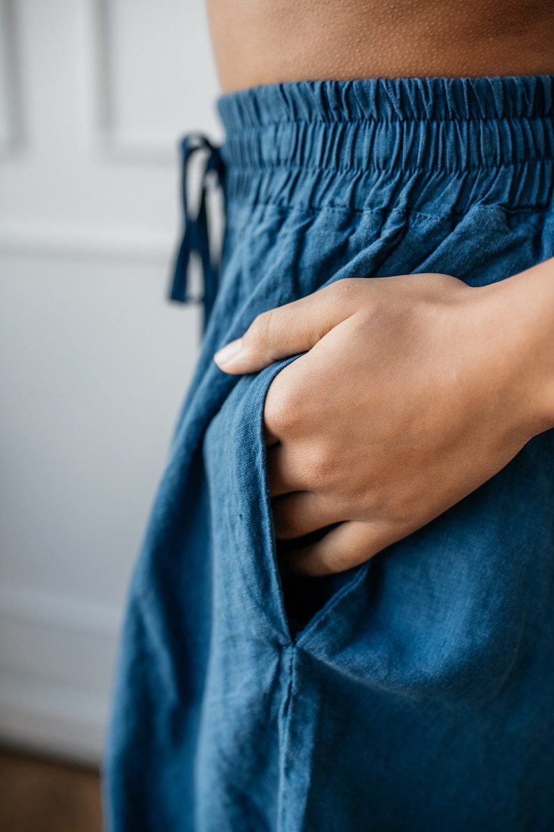 Pu&ograve; includere: Primo piano di pantaloni blu con elastico in vita e coulisse. Una mano &egrave; infilata nella tasca dei pantaloni, realizzati in un tessuto morbido e fluido. I pantaloni sembrano essere abbigliamento casual.