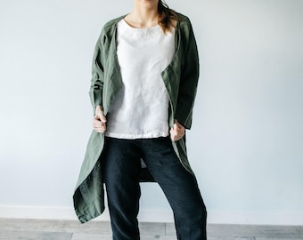 Può includere: Un cappotto lungo aperto color verde oliva indossato sopra un top bianco a maniche corte e pantaloni neri alla caviglia, abbinato a sneakers bianche con suola spessa. L'insieme presenta un look a strati con una giacca leggera.